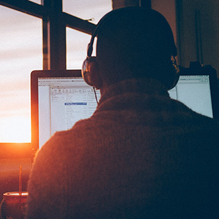A person wearing large headphones work at a computer while the sun sets